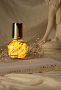 Ein Produktfoto eines golden leuchtenden Parfums, welches durch einen cremefarbenen Leinentuch Hintergund, dekorative Blumen und eine griechische Statue unterstützt wird.
